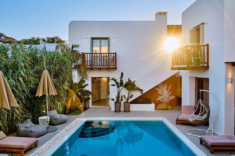 Dziedziniec z basenem, leżakami i zielenią przed białym budynkiem.