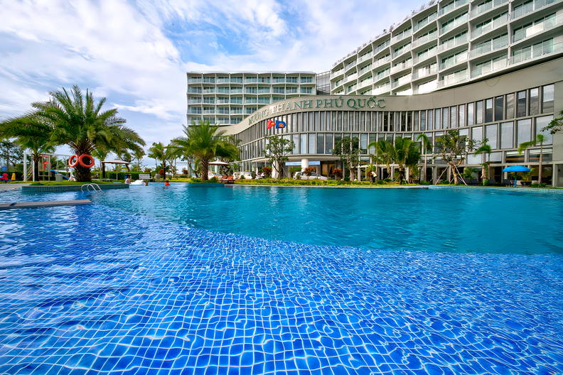 Basen z turkusową wodą przed hotelem otoczonym palmami.