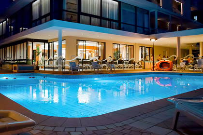 Nocny widok na oświetlony basen hotelowy i restaurację.