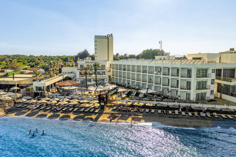 Widok z lotu ptaka na duży hotel i piaszczystą plażę z rzędami leżaków i parasoli nad błękitnym morzem.