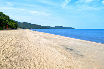 Szeroka, piaszczysta plaża nad błękitnym morzem, z zalesionymi wzgórzami w tle.