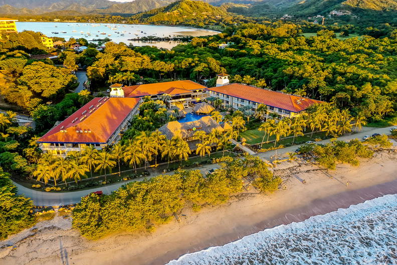 Hotel z basenem, palmami i plażą widziany z góry.