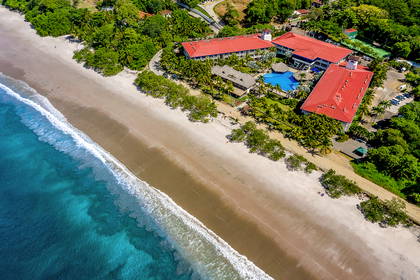 Hotel z czerwonym dachem, basenem i błękitnym oceanem widziany z lotu ptaka.