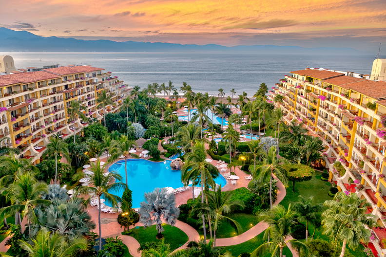 Widok z lotu ptaka na luksusowy kurort z palmami i basenami, w tle morze.