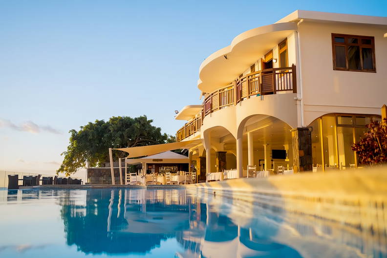 Błękitny basen odbija biały budynek z balkonami i restauracją.