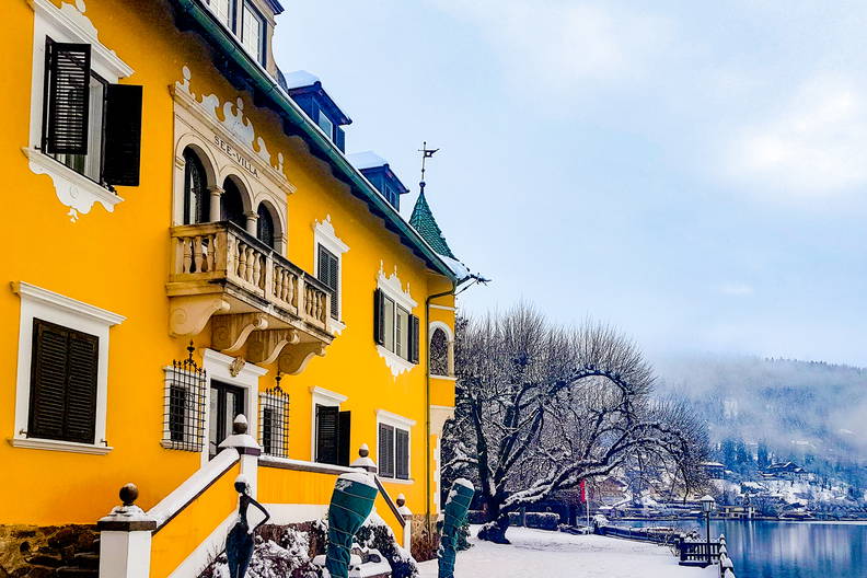 Żółty budynek z balkonem, ośnieżony, z widokiem na jezioro i wzgórza.
