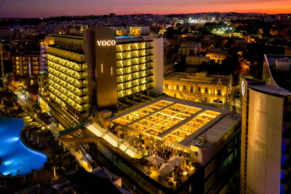 Nocny widok na oświetlony hotel VOCO z basenem i restauracją na dachu.