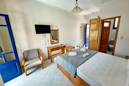 Jasny pokój hotelowy z podwójnym łóżkiem, drewnianymi meblami i otwartymi drzwiami do łazienki.