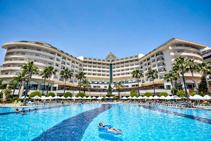 Hotel z basenem otoczonym palmami. Wokół relaksują się ludzie.