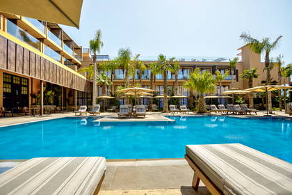 Basen hotelowy otoczony leżakami i palmami, z budynkiem hotelu w tle.