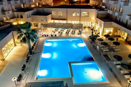 Hotelowy dziedziniec nocą z dużym, podświetlonym na niebiesko basenem, palmą i budynkami w tle.