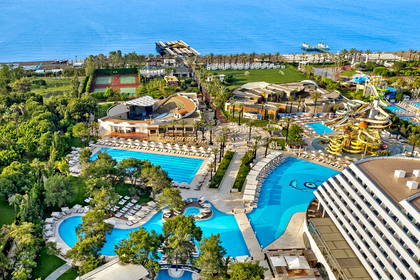 Widok z lotu ptaka na rozległy kompleks hotelowy nad morzem. Widać duże, błękitne baseny, aquapark i tereny zielone.