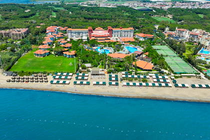 Widok z góry na rozległy kompleks hotelowy nad błękitnym morzem, z basenami, kortami tenisowymi i piaszczystą plażą.