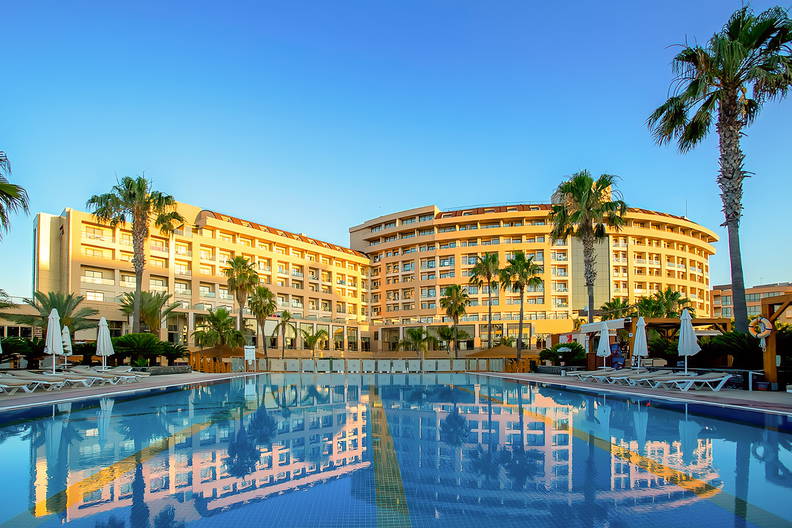 Duży hotel z basenem i palmami, którego fasada odbija się w wodzie.