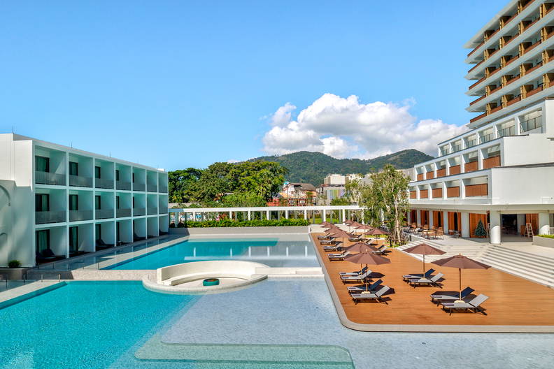 Nowoczesny hotel z basenem i drewnianym tarasem z leżakami i parasolami na tle zielonych wzgórz.