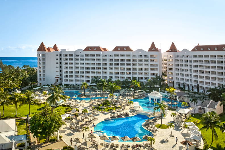 Rozległy, biały hotel z kompleksem basenów i palmami nad brzegiem oceanu w słoneczny dzień.
