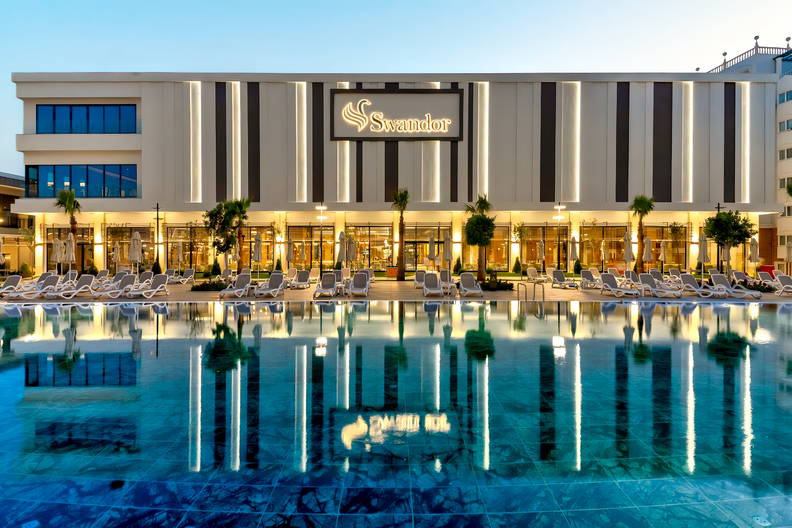 Nowoczesny, oświetlony hotel Swandor z basenem i leżakami o zmierzchu.