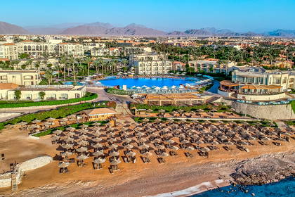 Widok z góry na resort z basenem i piaszczystą plażą z parasolami, z miastem i pasmem górskim w tle.