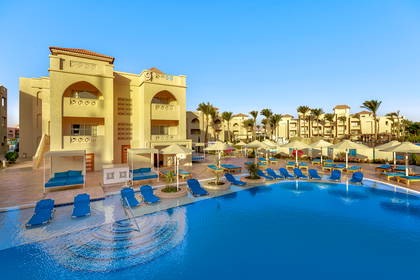Basen hotelowy z niebieskimi leżakami i palmami. W tle beżowe budynki resortu.