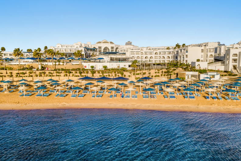 Biały hotel nad błękitnym morzem, z piaszczystą plażą pełną parasoli i leżaków.