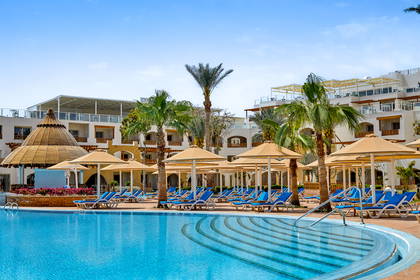 Hotelowy basen otoczony palmami, leżakami i parasolami, z budynkiem kurortu w tle.