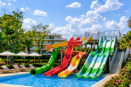 Aquapark z kolorowymi zjeżdżalniami wodnymi, palmami i leżakami wokół basenu.