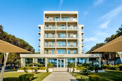 Nowoczesny, kilkupiętrowy hotel z balkonami, otoczony zadbanym trawnikiem i drzewami.