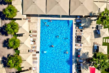 Widok z lotu ptaka na hotelowy basen z pływającymi ludźmi, otoczony leżakami, parasolami i palmami.