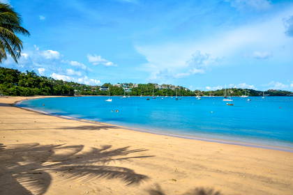 Piaszczysta plaża nad turkusową zatoką, po której pływają żaglówki. W tle zielone wzgórza i błękitne niebo z chmurami.