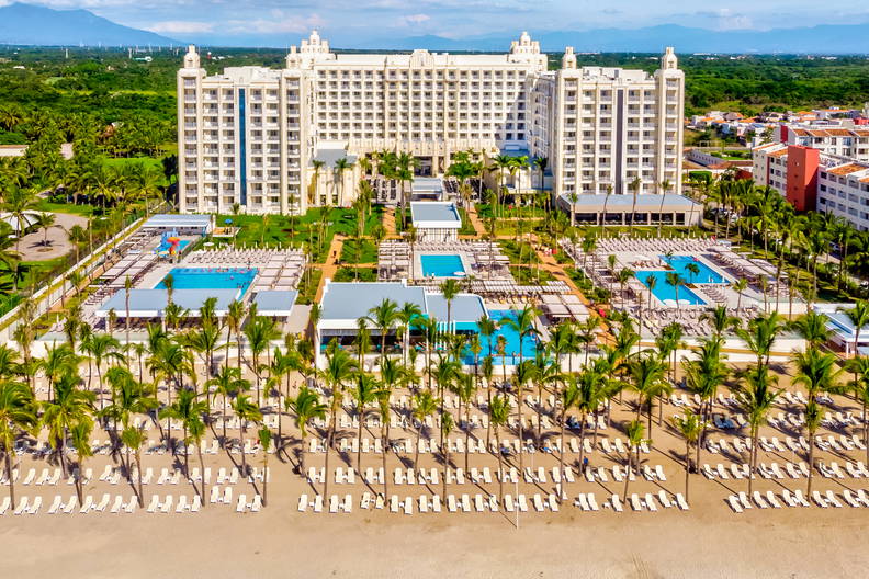 Widok z lotu ptaka na duży hotel przy plaży z basenami, palmami i leżakami ustawionymi w rzędach na piasku.