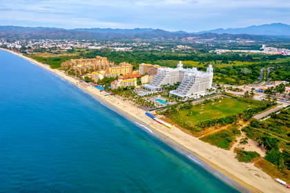 Widok z lotu ptaka na nadmorski kurort z hotelami, piaszczystą plażą i turkusowym oceanem. W tle widać góry.