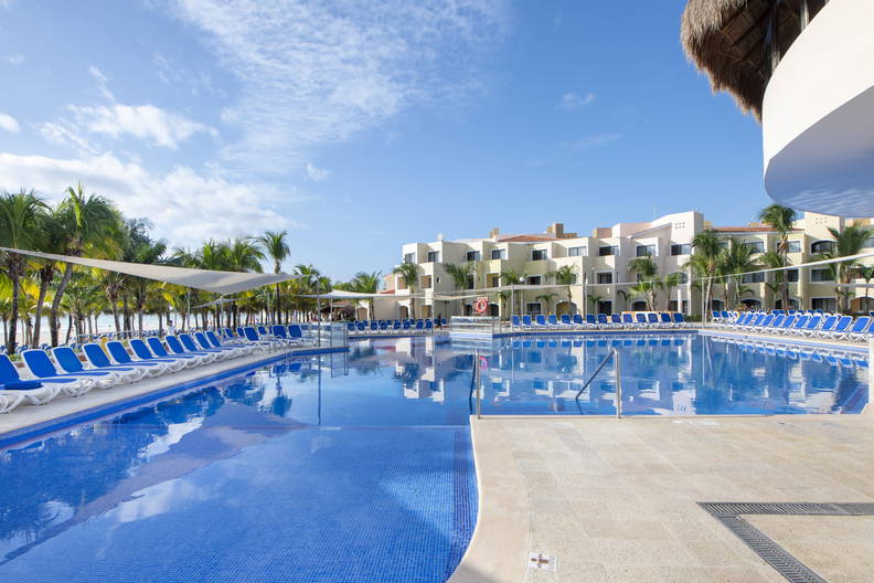 Duży basen hotelowy otoczony palmami i niebieskimi leżakami.