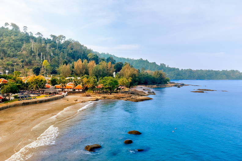 Widok z góry na zatokę z piaszczystą plażą, kurortem i zalesionym wzgórzem.