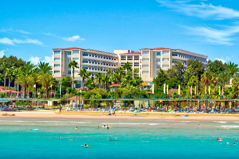 Piaszczysta plaża z palmami, duży hotel i ludzie kąpiący się w turkusowym morzu.