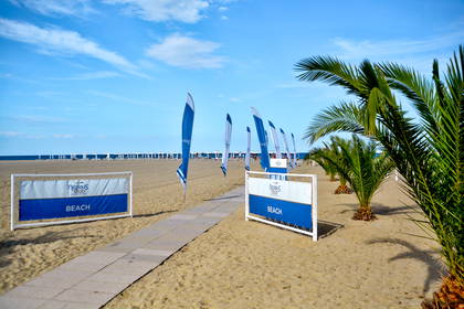 Drewniana ścieżka na piaszczystej plaży prowadzi do rzędów leżaków. Wejście na nią zdobią niebiesko-białe flagi i małe palmy.