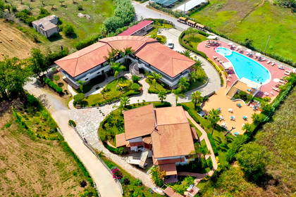 Kompleks hotelowy z basenem, otoczony zielenią i polami, sfotografowany z lotu ptaka.