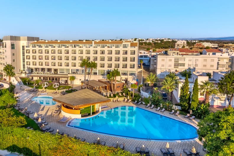 Ośrodek hotelowy z dużym, błękitnym basenem, barem i leżakami w otoczeniu zieleni.