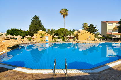 Duży hotelowy basen z błękitną wodą, otoczony leżakami, parasolami i palmami w słoneczny dzień.