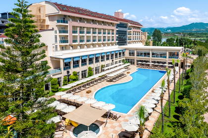 Nowoczesny hotel z dużym basenem otoczonym leżakami i zielenią, z górami w tle.