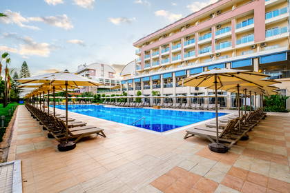 Duży basen hotelowy otoczony rzędami leżaków i parasoli, z budynkiem hotelu w tle.