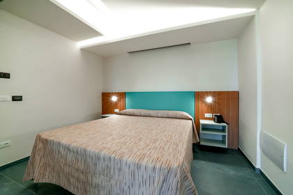 Nowoczesny pokój hotelowy z podwójnym łóżkiem, drewnianym zagłówkiem z turkusowym panelem i ciemną, kafelkową podłogą.