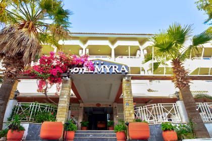 Główne wejście do Hotelu Myra w słoneczny dzień, otoczone palmami i różową bugenwillą.