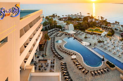 Widok z lotu ptaka na hotel The Sunset Bay z basenem, leżakami i morzem w tle podczas zachodu słońca.