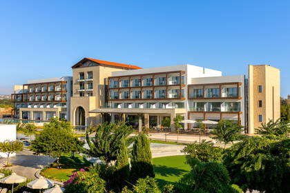 Nowoczesny wielopiętrowy hotel z kamienną elewacją i balkonami, otoczony ogrodem w słoneczny dzień.