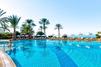 Hotelowy basen z błękitną wodą, palmami i rzędem leżaków przy plaży.