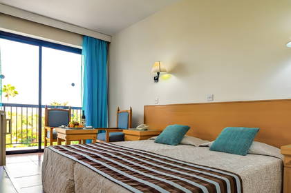 Jasny pokój hotelowy z łóżkiem, niebieskimi zasłonami, balkonem oraz stolikiem z owocami.