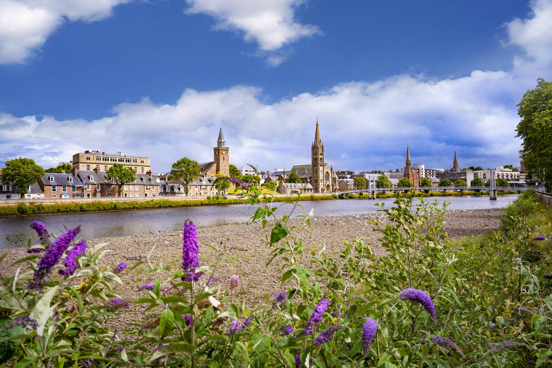 Słoneczny widok na miasto Inverness i rzekę Ness, z fioletowymi kwiatami na pierwszym planie i wieżami kościołów w tle.