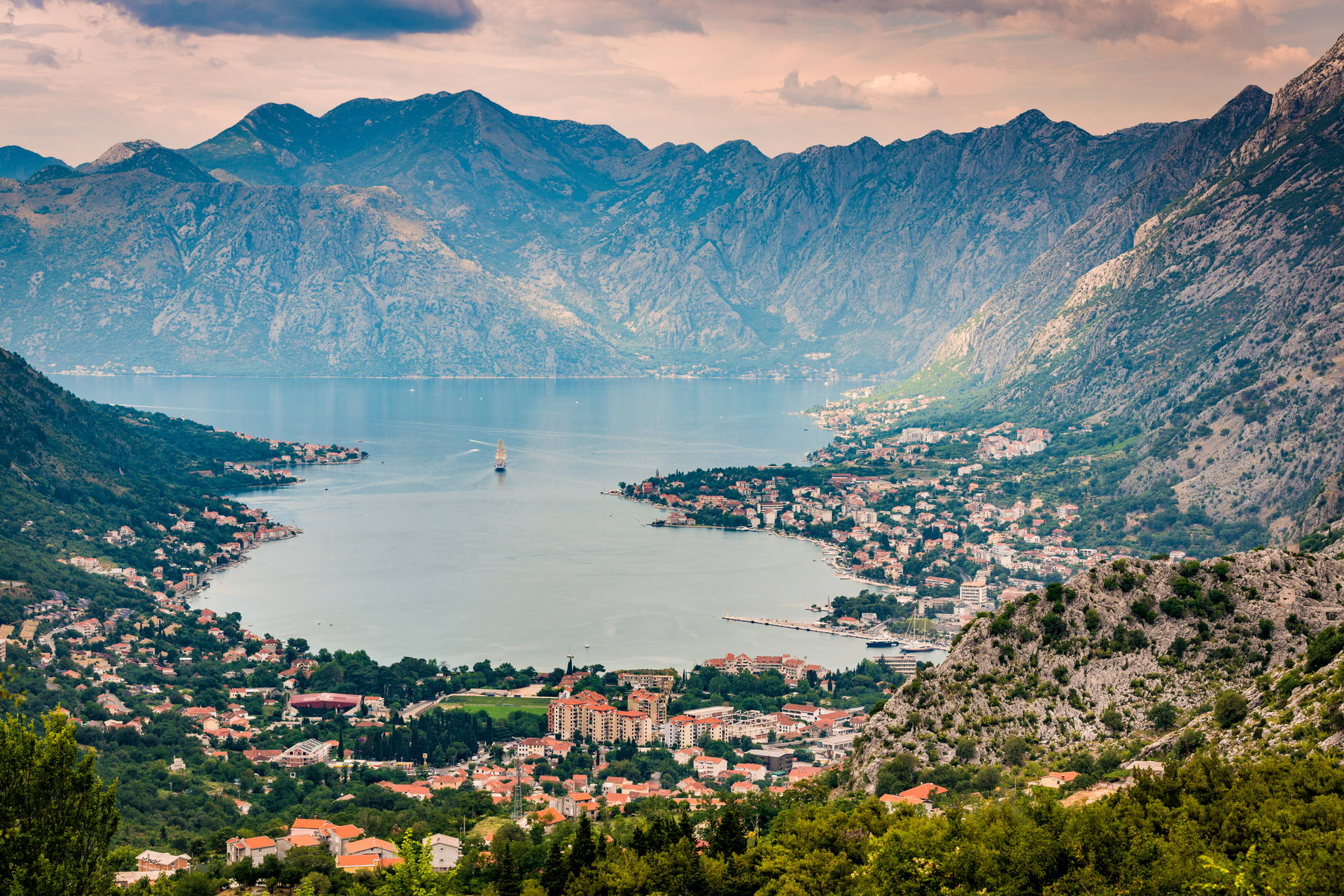 Boka Kotorska z górami w tle, miastem u wybrzeża i żaglówką na wodzie.