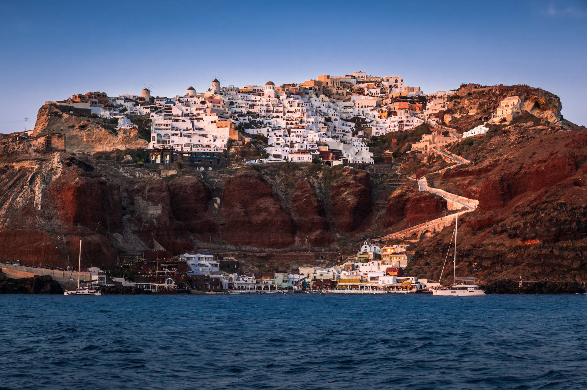 Widok na białe miasteczko Oia na klifie Santorini, łodzie na morzu i zachód słońca.