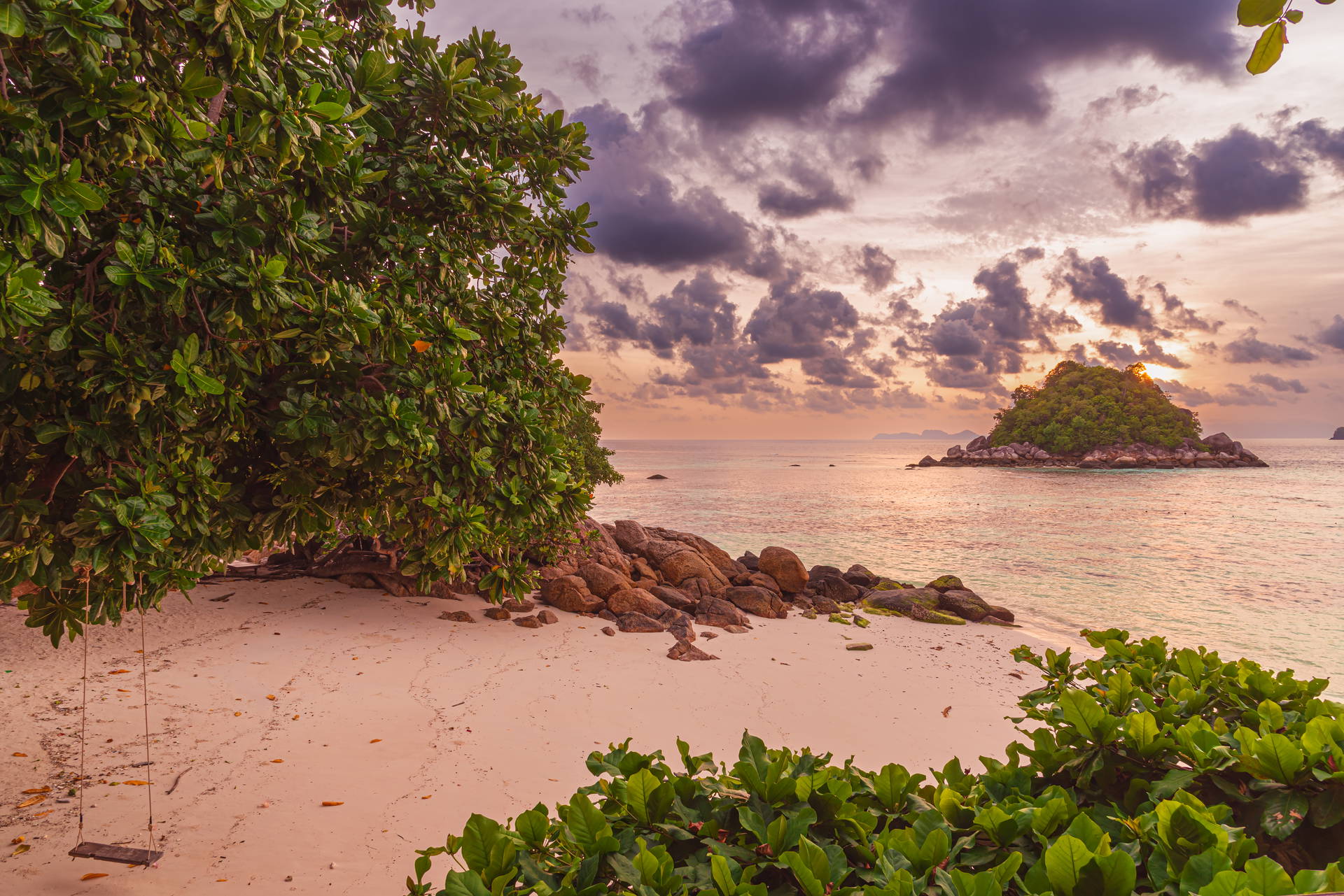 Piaszczysta plaża na wyspie Koh Lipe w Tajlandii z huśtawką na drzewie, skałami i widokiem na wysepkę podczas zachodu słońca.